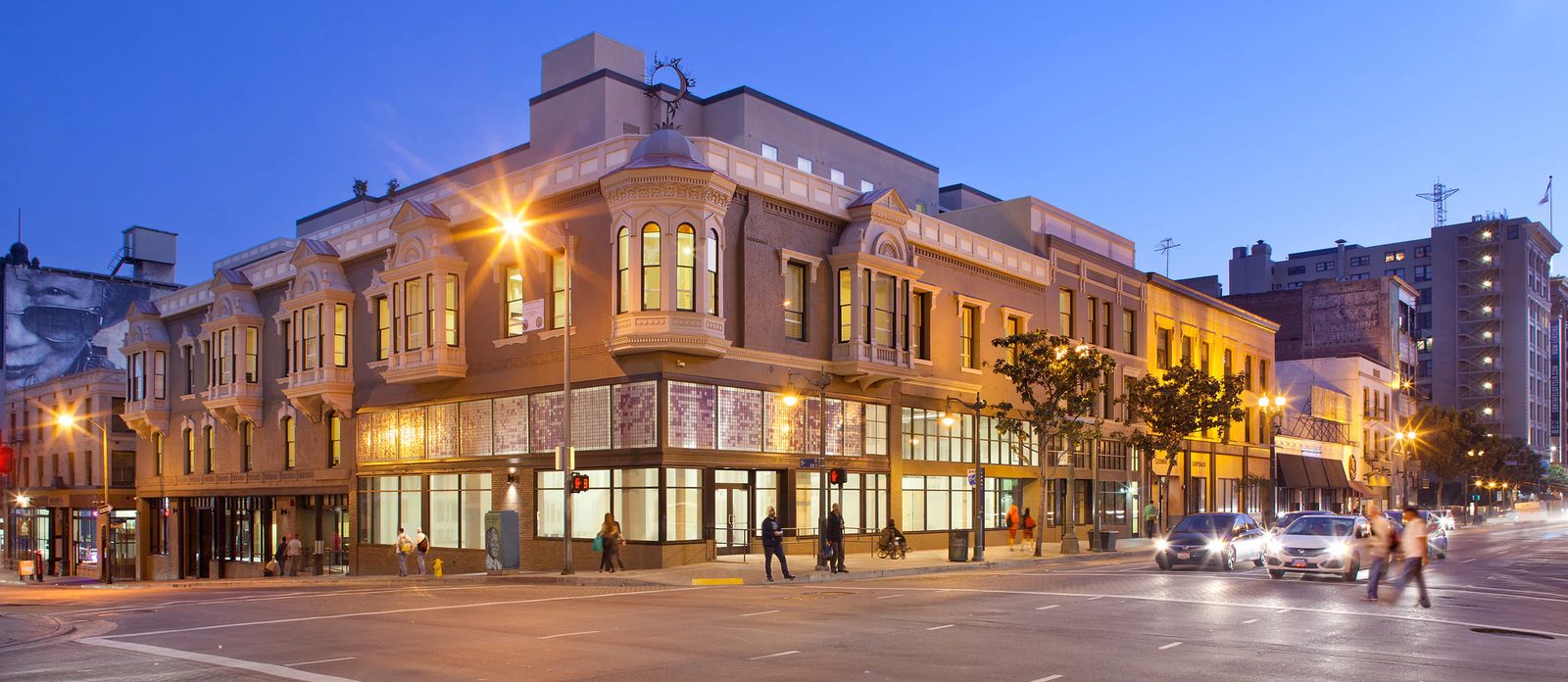 A restored historic brick building with ornate bay windows on a downtown Los Angeles street corner at dusk, its ground-floor windows lit